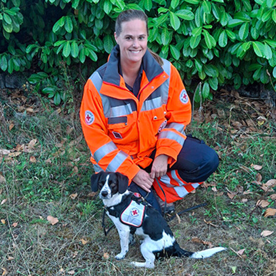 Eine Frau kniet in Einsatzbekleidung, vor ihr sitzt ein Hund.