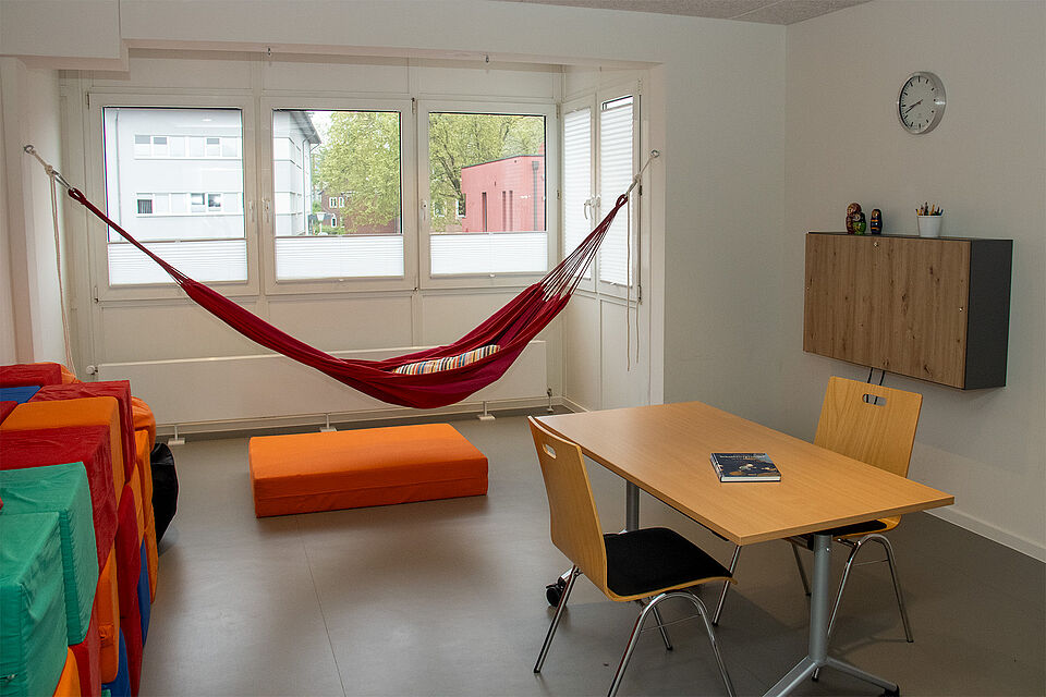 Raum mit Fenster, einem Tisch und zwei Stühlen, einer Hängematte und zahlreichen Kissen.