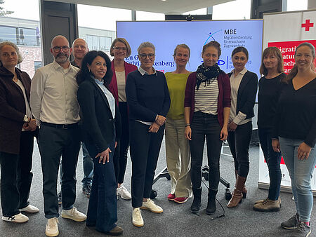 Eine Gruppe von elf Personen steht in einem Innenraum vor zwei Informationsbannern und einem Bildschirm.