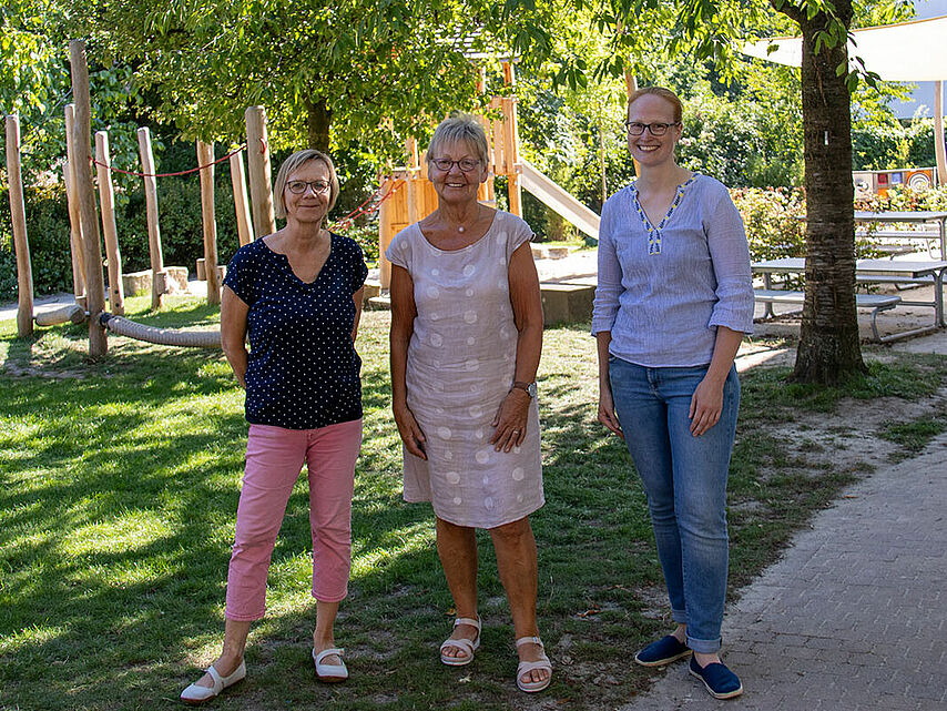 Drei Personen stehen im Freien in einem Park oder auf einem Spielplatz. Im Hintergrund sind Bäume, Rasenflächen und Spielgeräte zu sehen.