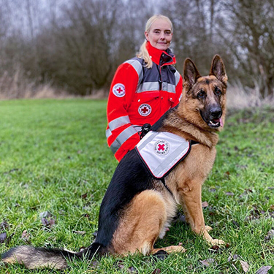 Eine Person in Einsatzkleidung kniet auf einer Wiese. Vor ihr sitzt ein Hund, der eine Weste mit dem Rotkreuz-Logo und dem Aufdruck „Rettung“ trägt. Im Hintergrund sind Bäume zu sehen.