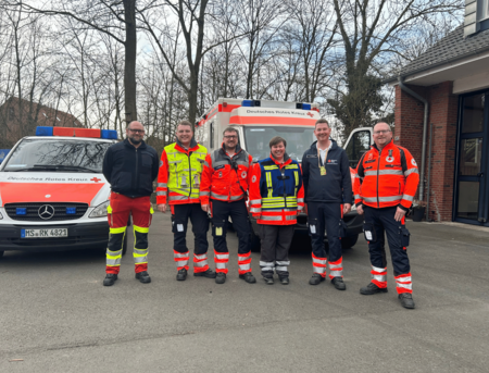 Sechs Personen in Einsatzkleidung vor einem Krankentransportwagen und einem Notarzteinsatzfahrzeug des DRK