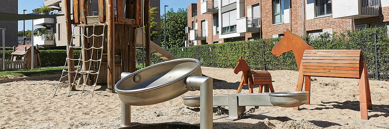 Erphobogen Slider Ein Spielplatz mit Sand, einem großen Klettergerüst, zwei Holzpferden und Wasserspielen. Im Hintergrund Wohnhäuser.