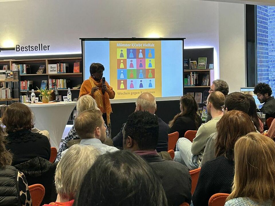 Eine Frau mit Mikro steht vor einer Gruppe, im Hintergrund befinden sich Bücherregale
