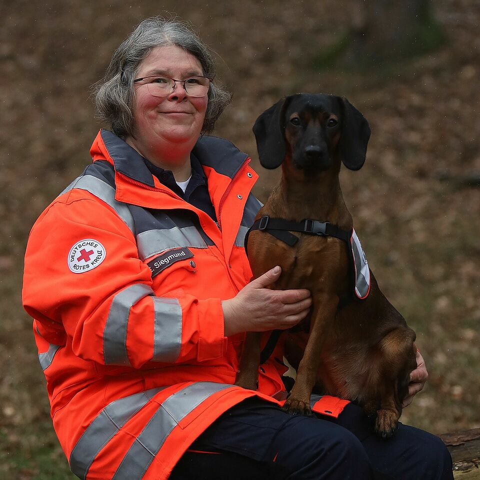 Eine Person in Einsatzkleidung hält einen braunen Hund im Arm, der ein Geschirr trägt. Auf dem Ärmel der Kleidung ist das Logo des Deutschen Roten Kreuzes zu sehen.