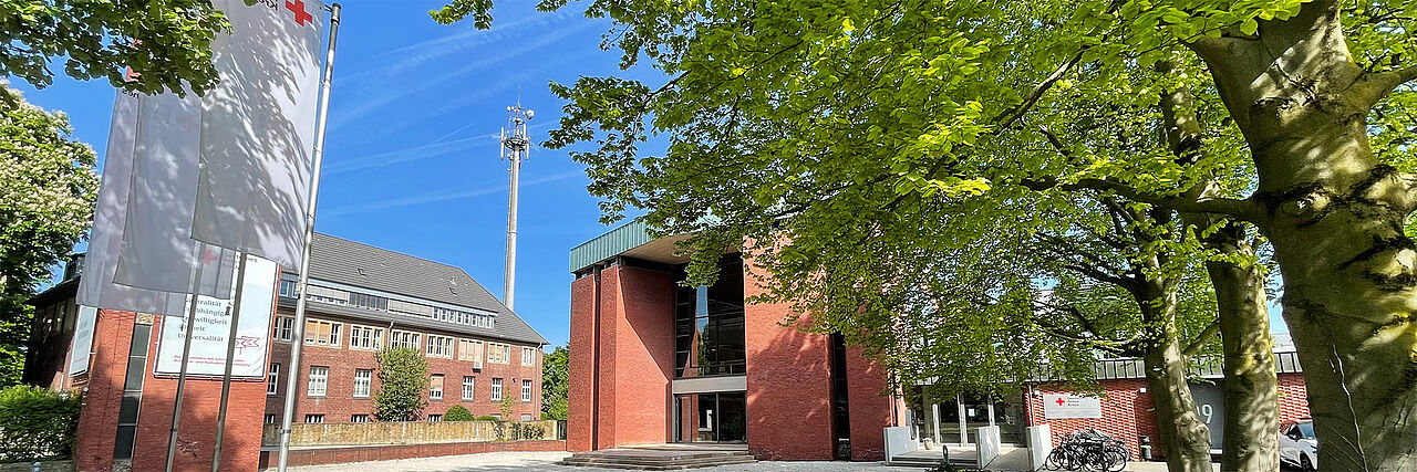 Cheruskerring April Ein Gebäude aus roten Ziegelsteinen steht im Zentrum des Bildes. Es ist von Bäumen und grünen Pflanzen umgeben. Links im Bild ragt ein Fahnenmast mit drei weißen Flaggen empor, auf der ein rotes Kreuz zu sehen ist. Im Hintergrund befindet sich ein weiteres Gebäude mit traditioneller Architektur und vielen Fenstern. Der Himmel ist klar und blau.