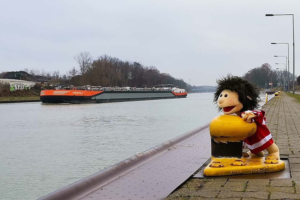 Ein breiter Kanal mit einem großen Frachtschiff, das auf dem Wasser fährt. Rechts verläuft ein gepflasterter Weg mit Straßenlaternen und einem gelben Poller. Daneben steht eine kleine Figur in rot-weiß gestreifter Kleidung. Im Hintergrund sind Bäume und einige Gebäude am linken Ufer zu sehen.
