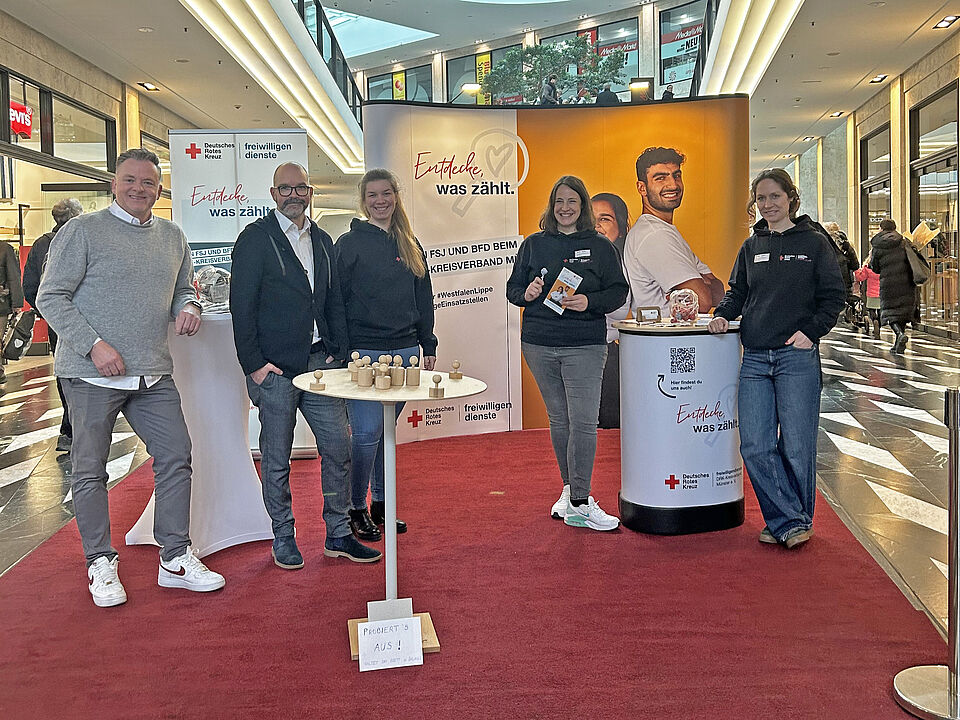 Fünf Personen stehen an einem Stand im Einkaufszentrum auf einem roten Tepich. Im Hintergrund sind Aufsteller mit Informationen zum Freiwilligendienst.