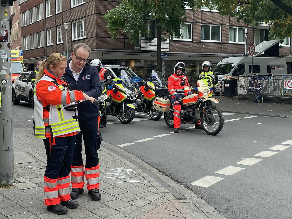Giro 2025 Zwei Einsatzkräfte besprechen sich neben einer Straße, auf der Motorräder in Einsatzkleidung warten