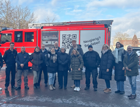 Eine Gruppe von Menschen steht auf einem Innenhof vor einem Feuerwehrauto. Der Boden ist vereist, im Hintergrund sieht man Bäume und blauen Himmel.