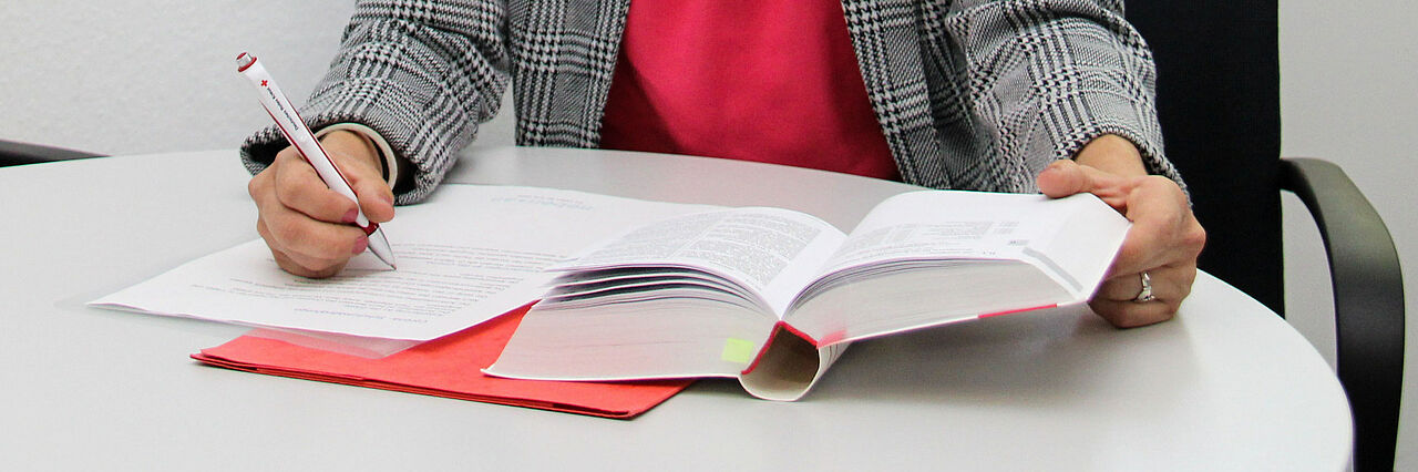 Eine Person sitzt an einem Schreibtisch mit einem Gesetzesbuch, einem Blatt Papier und einem Stift