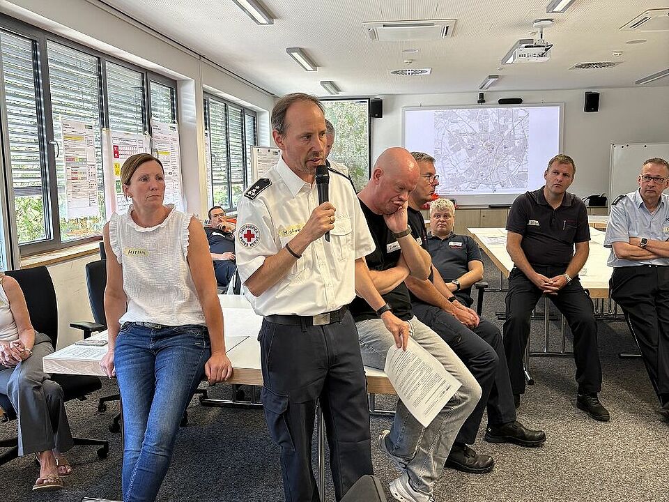 Mehrere Personen stehen in einem Halbkreis in einem Raum und hören einer Person mit Mikrofon zu. Im Hintergrund sind eine Lagekarte und Plakate zu sehen.