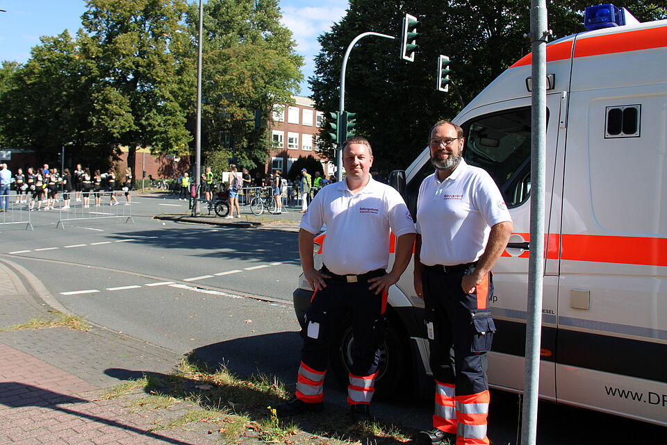 Marathon Zwei Personen in Einsatzkleidung neben einem Einsatzfahrzeug, im Hintergrund Laufstrecke des Marathons