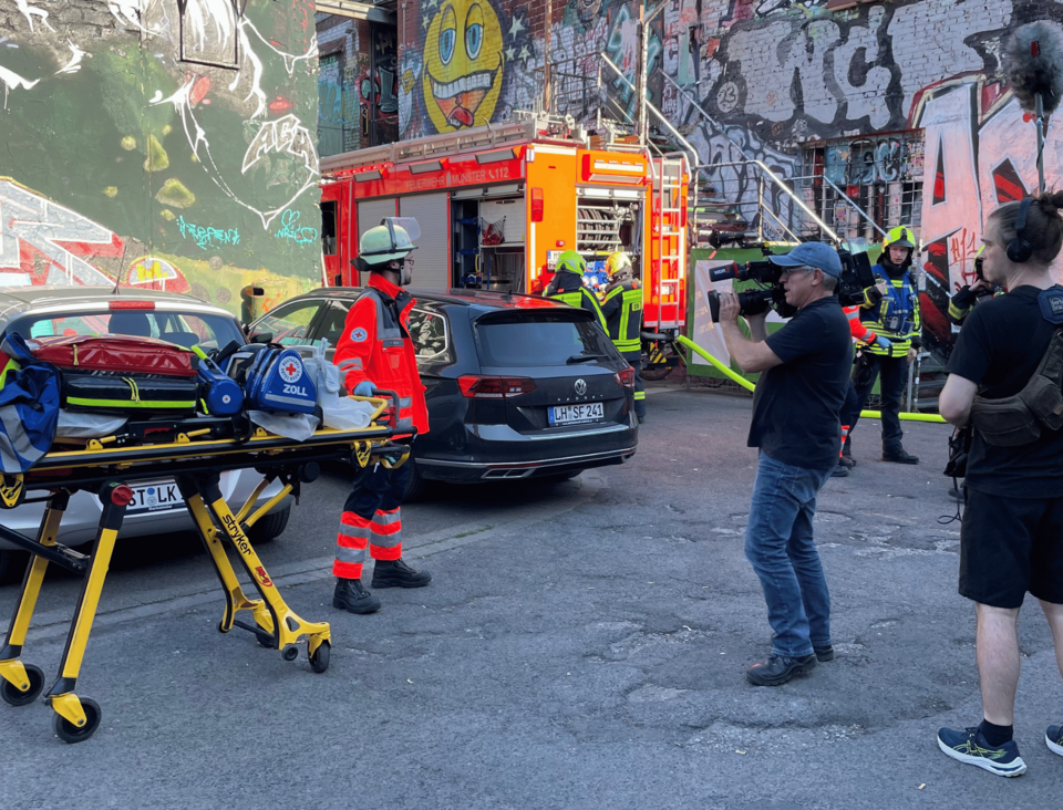 Eine Person in Einsatzkleidung schiebt eine Trage mit Rettungsequipment und wird dabei von einem Kamerateam gefilmt. Im Hintergrund ist ein Feuerwehreinsatzauto und weitere Einsatzkräfte zu sehen.