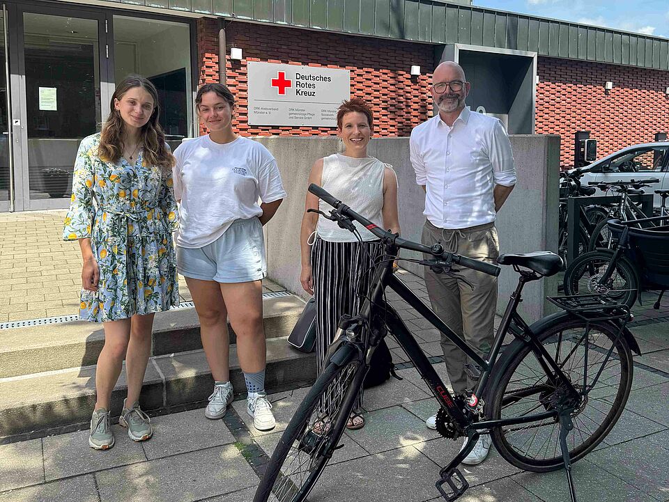 Vier Personen mit einem Fahrrad vor einem Gebäude mit DRK-Logo