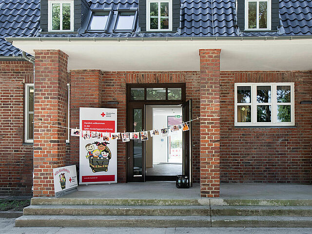 Eingang eines Backsteingebäudes mit Ziegeldach und zwei großen Säulen, die das Vordach stützen. Drei Stufen führen zur Eingangstür hinauf. Über dem Eingang hängt eine Girlande mit Fotos. Links neben der Tür steht ein Aufsteller. Daneben befindet sich ein weiteres Schild auf dem Boden. Durch die geöffnete Tür ist der Innenraum des Gebäudes sichtbar.