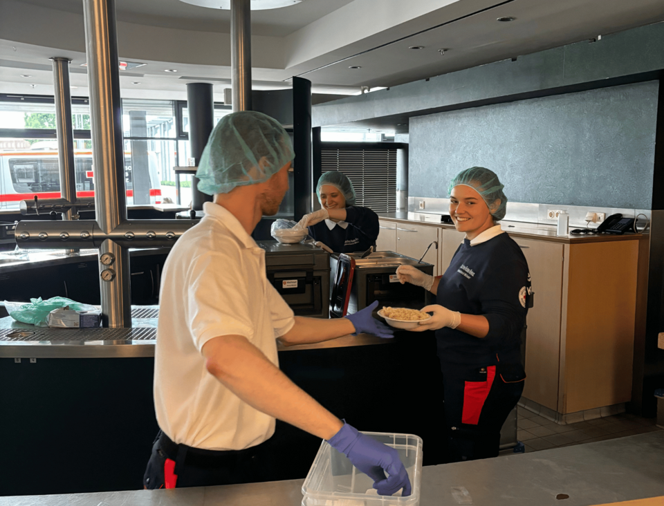 Drei Personen in Einsatzkleidung und Haarnetzen stehen in einer Küche und servieren Essen