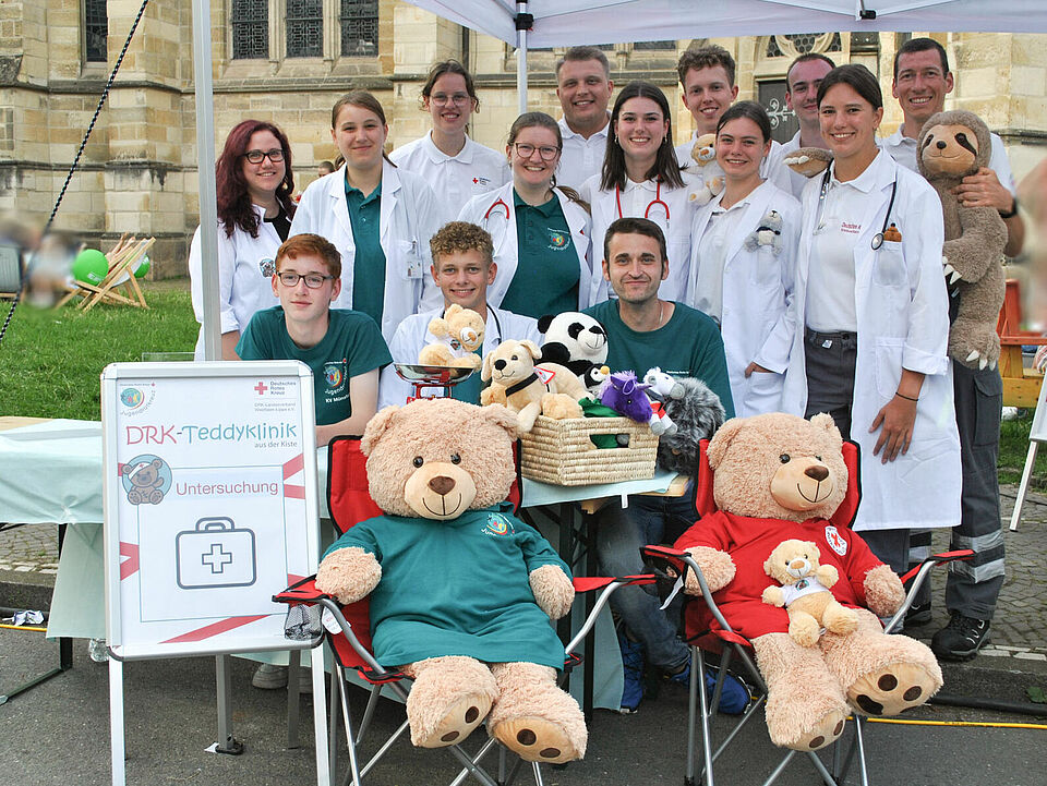 Ein Gruppenbild der Teddyklinik unter einem weißen Pavillion. Im Vordergrund ist ein Schild der Teddyklinik und mehrere Teddys zu sehen.