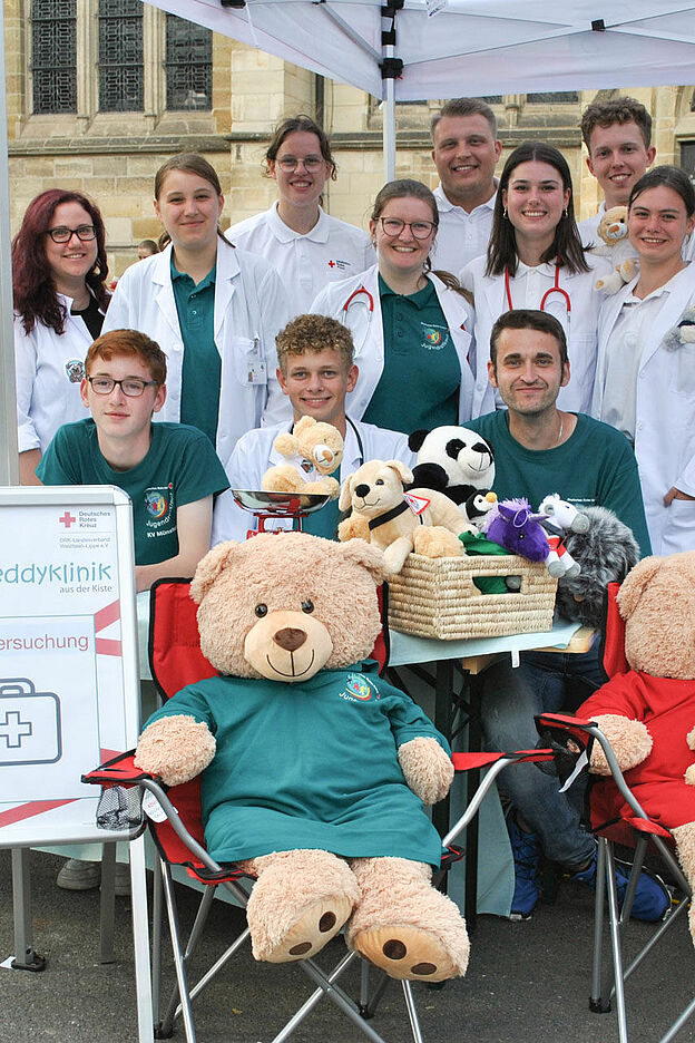 Ein Gruppenbild der Teddyklinik unter einem weißen Pavillion. Im Vordergrund ist ein Schild der Teddyklinik und mehrere Teddys zu sehen.