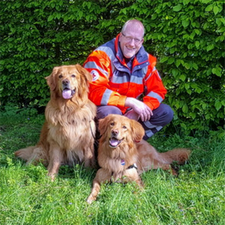 Eine Person in Einsatzkleidung kniet neben zwei Hunden. Im Hintergrund befindet sich grüne Vegetation.