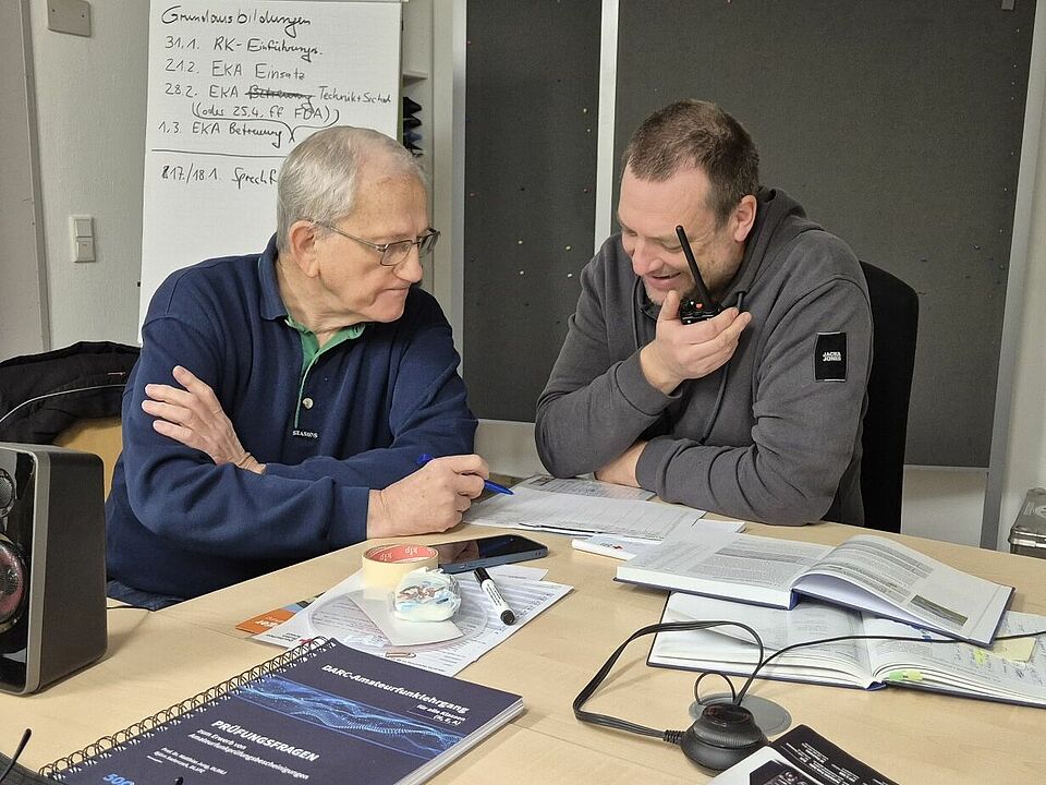 Amateurfunk Zwei Personen sitzen an einem Tisch, vor ihnen liegen Bücher und weitere Unterlagen. Eine Person hält ein Funkgerät in der Hand.