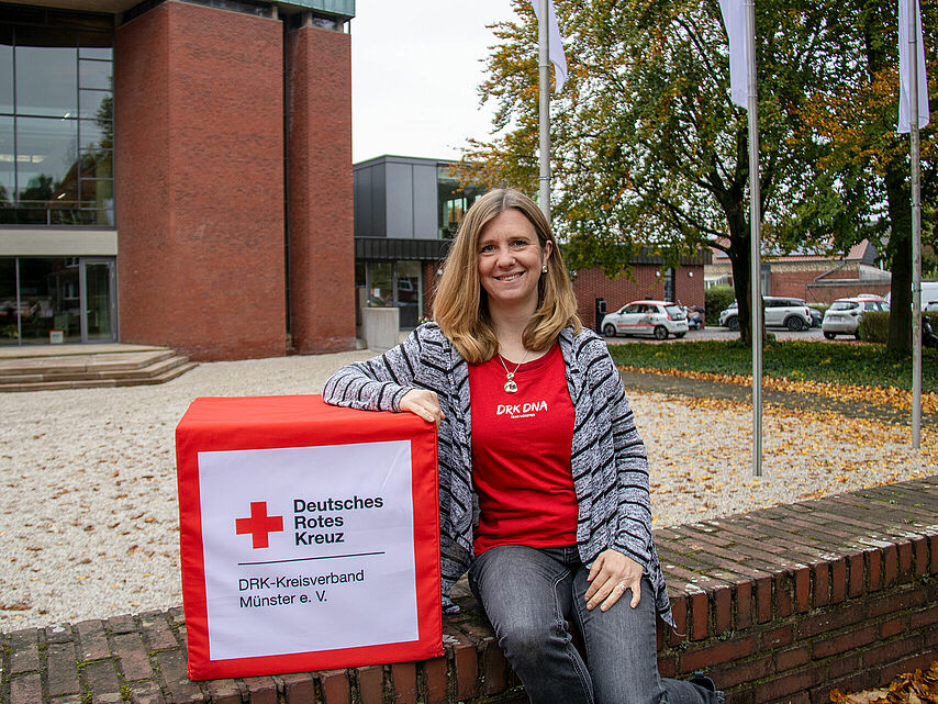 Eine Person sitzt auf einer gemauerten Kante neben einer Box mit der Aufschrift „Deutsches Rotes Kreuz DRK-Kreisverband Münster e. V.“. Die Person trägt ein rotes Shirt mit weißem „DRK DNA“-Schriftzug. Im Hintergrund sind ein modernes Gebäude mit großen Fenstern, Bäume und parkende Autos zu sehen.