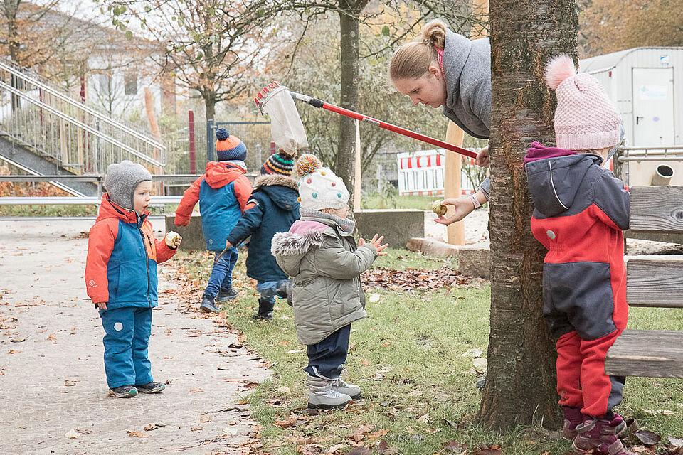Fünf Kinder in warmer Kleidung mit einer erwachsenen Person. Sie stehen neben einem Baum und halten einen Apfelpflücker in der Hand