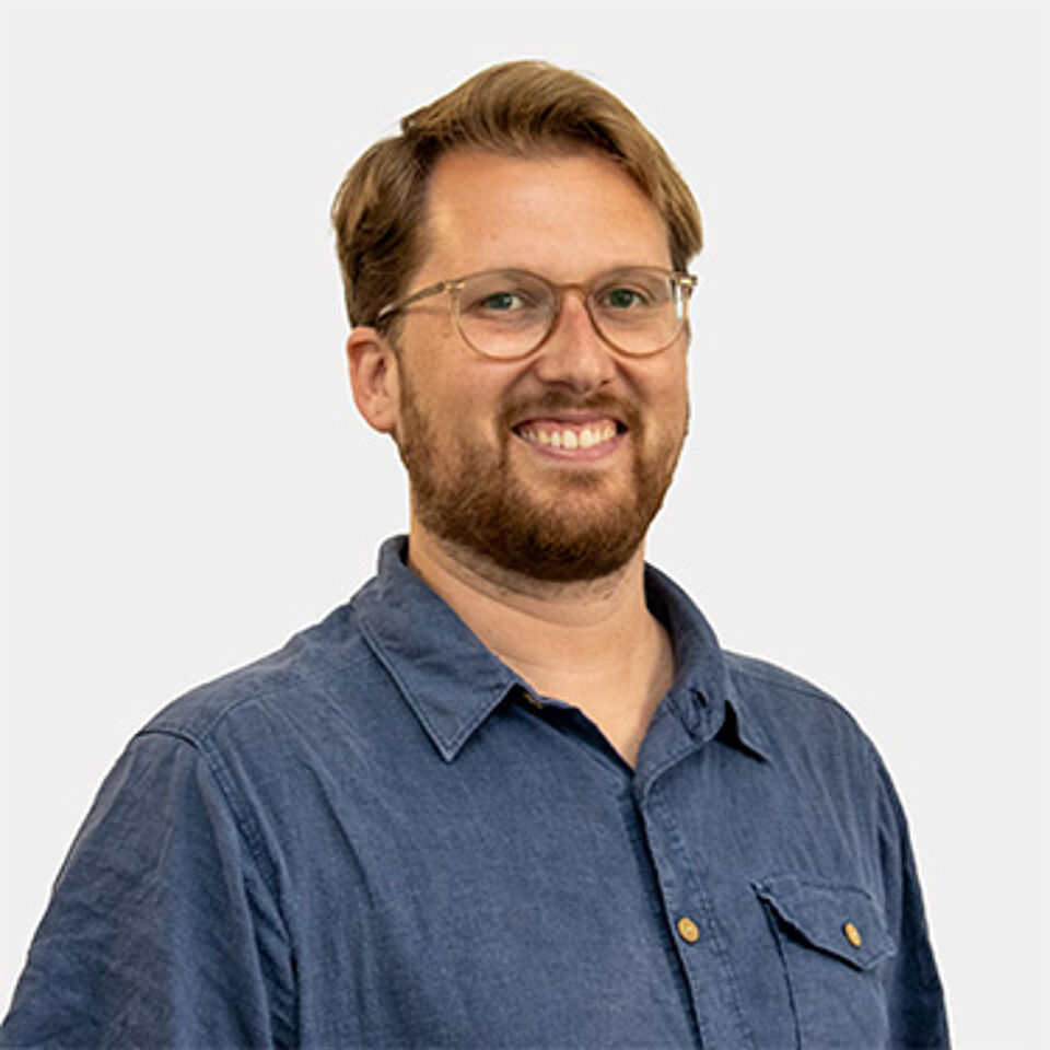 Mann mit blonden Haaren, Brille und blauem Hemd vor weißem Hintergrund