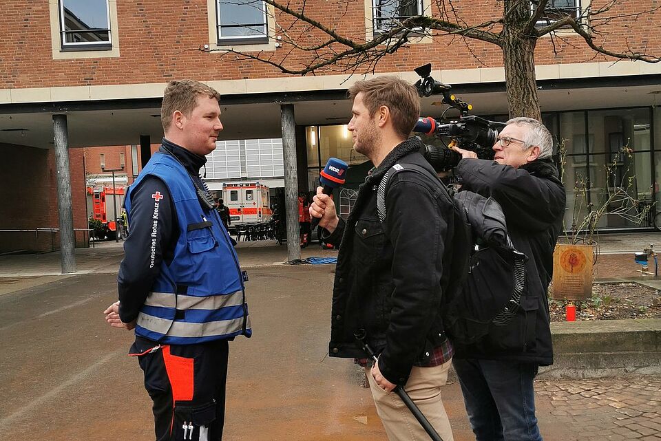 Rosenmontag Person in Einsatzkleidung und blauer Weste wird von einem Kamerateam interviewt