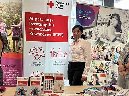 Beratungsmesse Eine Frau steht hinter einem Tisch mit Flyern und weiteren Materialien und neben einem Roll-Up mit dem DRK-Logo und "Migrationsberatung für erwachsene Zugewanderte"