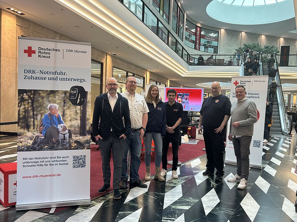 Sechs Personen stehen in einem Einkaufszentrum vor einem DRK-Stand. Links im Bild steht ein Roll-Up mit Informationen zum DRK-Hausnotruf.