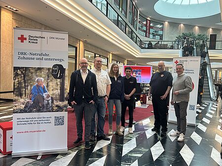 Sechs Personen stehen in einem Einkaufszentrum vor einem DRK-Stand. Links im Bild steht ein Roll-Up mit Informationen zum DRK-Hausnotruf.