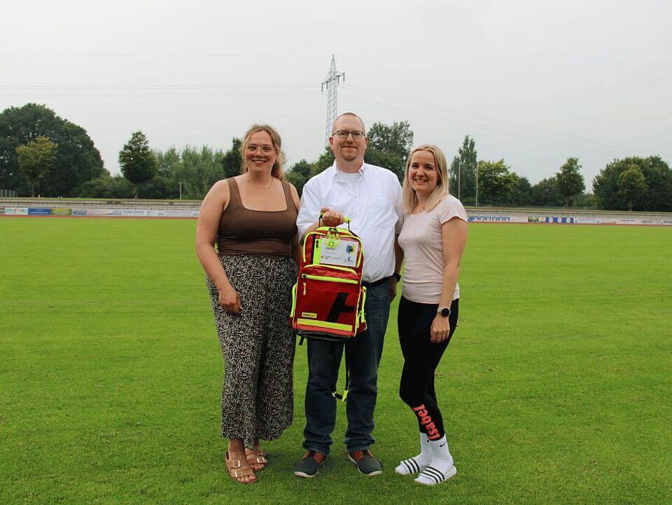 Übergabe Rucksack Drei Personen stehen auf einem Sportplatz und halten einen Notfallrucksack in die Kamera.