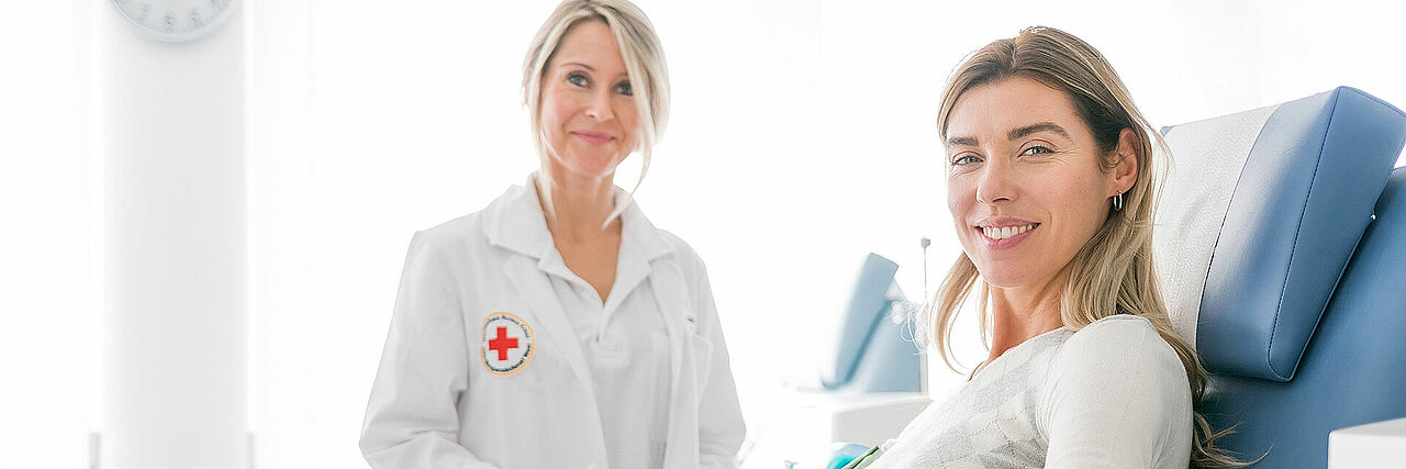 Zwei Personen befinden sich in einem medizinischen Umfeld. Eine der Personen trägt einen weißen Kittel mit einem Rotkreuz-Emblem auf der linken Seite, was auf eine Tätigkeit im Gesundheitswesen hinweist. Die andere Person sitzt und scheint medizinisch betreut oder beraten zu werden.