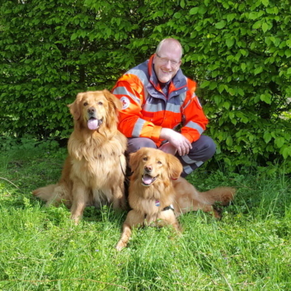 Eine Person in Einsatzkleidung kniet neben zwei Hunden. Im Hintergrund befindet sich grüne Vegetation.