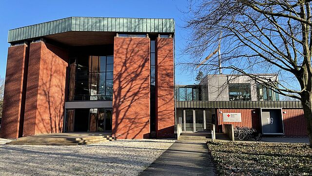 Ein rotes Backsteingebäude mit einer Fensterfront. Recht ist ein Anbau mit einem gläsernen Aufbau angebaut. Neben der Tür hängt ein Schild des DRK-Kreisverbandes Münster. Vor dem Gebäude ist ein Kiesfeld, ein Weg und Grün.