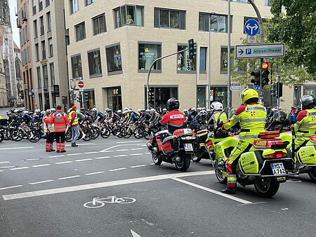 Rettungskräfte auf Motorrädern stehen in Position, auf der Straße ist ein Feld von Radfahrer*innen. Zwei Einsatzkräfte koordinieren die Situation. Im Hintergrund ist ein Kirchturm zu sehen.