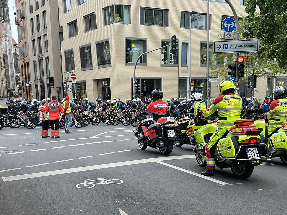 Giro 2025 Rettungskräfte auf Motorrädern stehen in Position, auf der Straße ist ein Feld von Radfahrer*innen. Zwei Einsatzkräfte koordinieren die Situation. Im Hintergrund ist ein Kirchturm zu sehen.