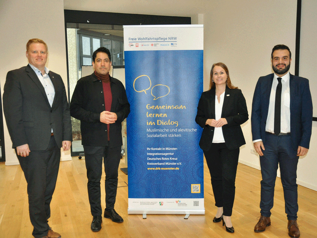 Vier Personen stehen vor einem Banner mit dem Titel: ‚Freie Wohlfahrtspflege NRW – Gemeinsam lernen im Dialog. Muslimische und alevitische Sozialarbeit stärken.‘