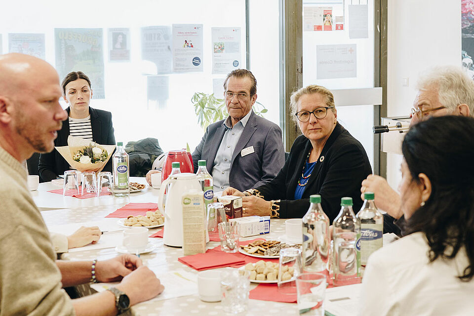 Besuch Svenja Schulze Eine Gruppe sitzt um einen Tisch mit Snacks und Getränken, im Hintergrund sind Plakate an den Fenstern zu sehen.