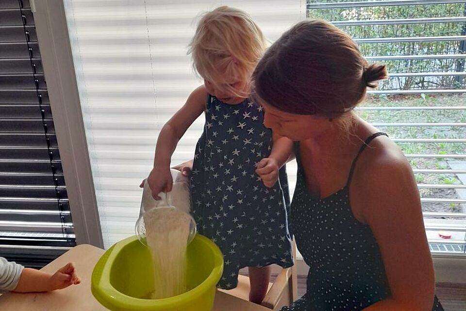 Ein Kind und eine erwachsene Person führen gemeinsam eine Aktivität durch. Das Kind schüttet eine weiße, pulverartige Substanz – vermutlich Mehl – aus einem durchsichtigen Behälter in eine große grüne Schüssel. Die erwachsene Person unterstützt und beaufsichtigt das Kind dabei. Sie befinden sich in einem Innenraum nahe einem Fenster mit teilweise geöffneten Jalousien, durch das Tageslicht einfällt. Am linken Bildrand ist eine weitere Person teilweise zu sehen