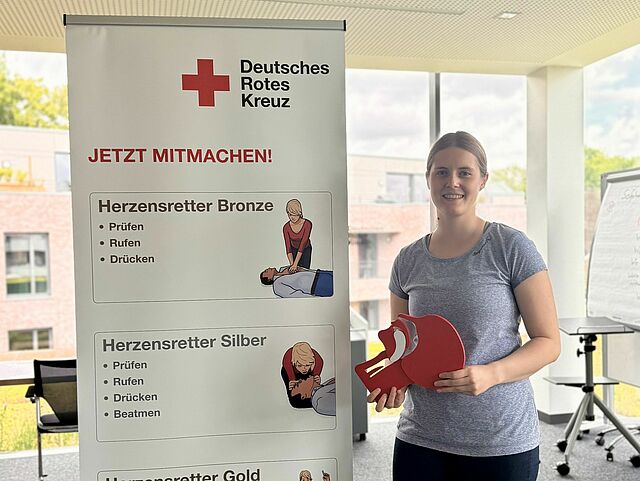In einem hellen Schulungsraum steht eine junge Frau und hält ein Schluckmodell n in der Hand. Neben ihr steht ein Roll-Up des Deutschen Roten Kreuzes mit dem Titel „Jetzt mitmachen“ und Informationen zu den drei Stufen des „Herzensretter“-Programms: Bronze, Silber und Gold