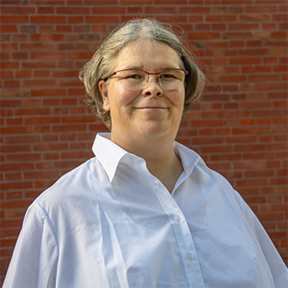 Frau mit grauen Haaren und Brille in einer weißen Bluse vor Backstein-Hintergrund