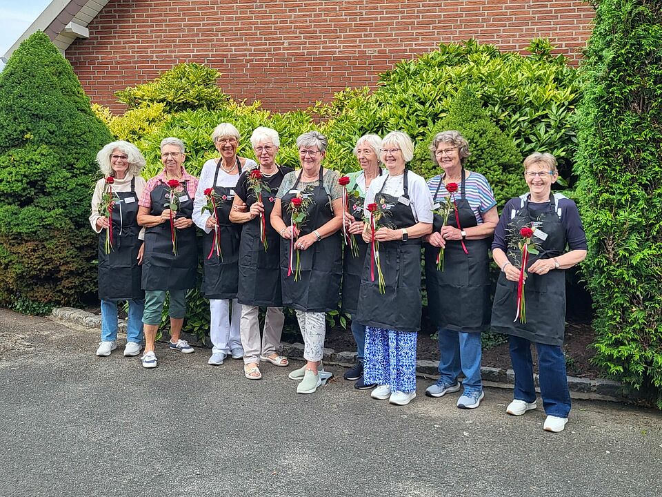 Neun Frauen mit Schürzen halten eine Rose in der Hand und stehen vor einer grünen Hecke, im Hintergrund ist ein Haus zu sehen