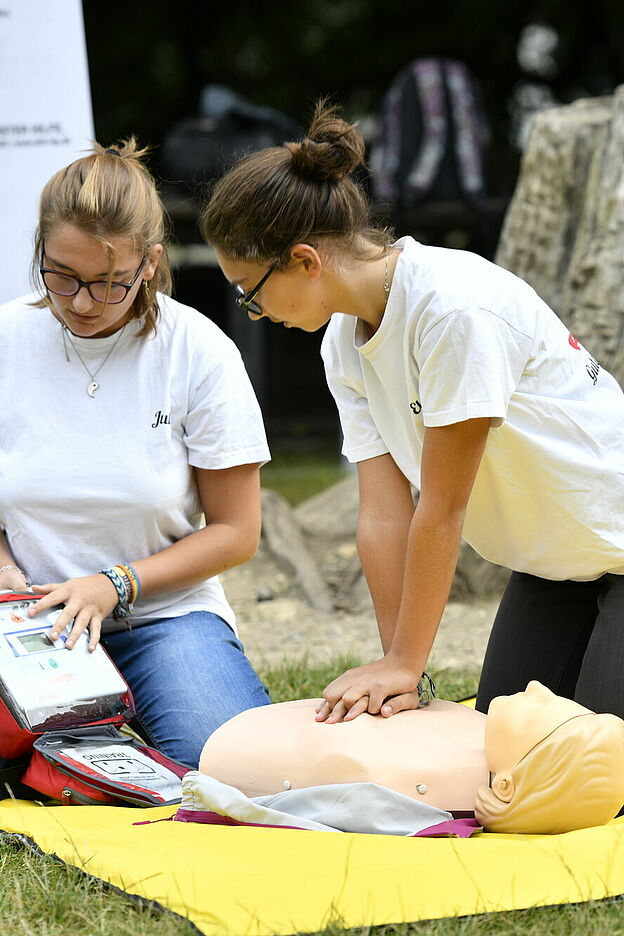 Im Freien findet ein Erste-Hilfe-Training statt. Eine Person führt auf einer gelben Matte Herzdruckmassagen an einer Reanimationspuppe durch. Eine zweite Person beobachtet und unterstützt möglicherweise mit medizinischem Equipment aus einem geöffneten Notfallkoffer. Im Hintergrund sitzt eine dritte Person in der Nähe eines Baumstumpfs, umgeben von weiteren medizinischen Materialien.