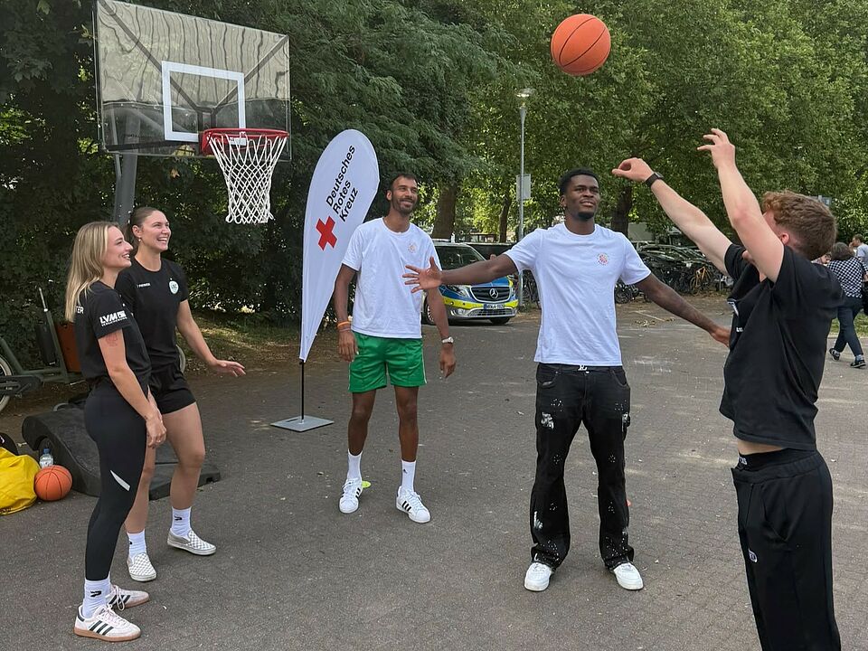 Fünf junge Menschen stehen vor einem Basketballkorb und werfen einen Basketball. Im Hintergrund ist eine DRK-Beachflag zu sehen.