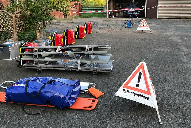 In einem Außenbereich sind mehrere Notfallrucksäcke in Rot und Gelb ordentlich nebeneinander aufgestellt. Daneben liegen gestapelte Tragen, ein orangefarbenes Spineboard mit einer blauen Tasche darauf sowie zwei Warndreiecke mit der Aufschrift „Patientenablage“. Im Hintergrund sind grüne Pflanzen, ein Backsteingebäude und eine Garage mit Absperrband zu sehen.