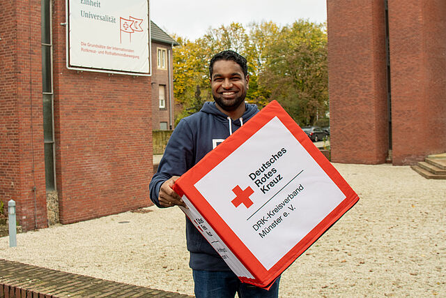 Eine Person hält eine große Box mit dem Logo und der Aufschrift „Deutsches Rotes Kreuz DRK-Kreisverband Münster e. V.“
