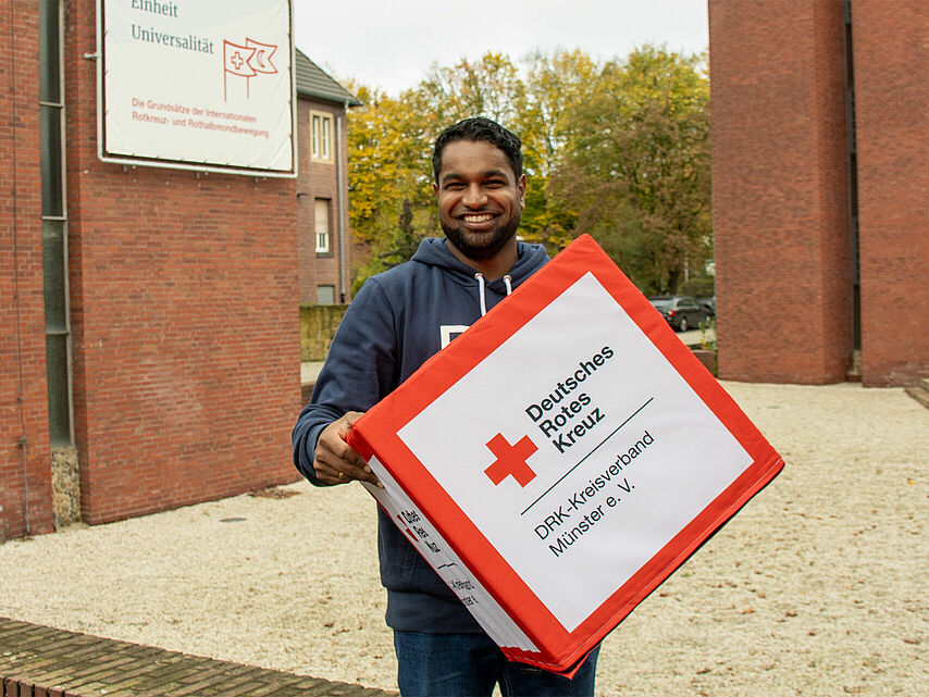 Eine Person hält eine große Box mit dem Logo und der Aufschrift „Deutsches Rotes Kreuz DRK-Kreisverband Münster e. V.“