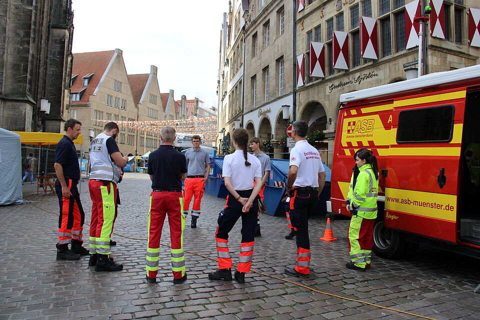 Einsatzbesprechung Eine Gruppe von Einsatzkräften steht in einem Kreis, im Hintergrund Giebelhäuser mit Wimpeln